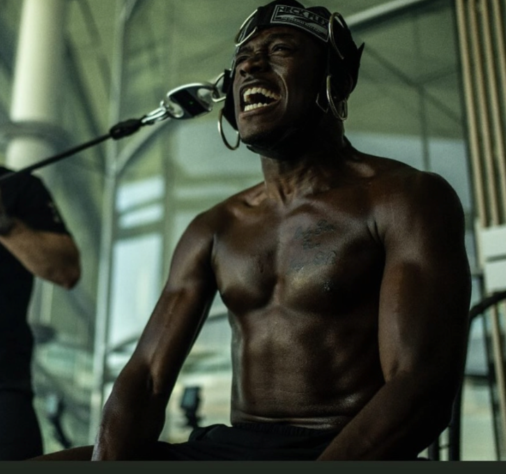 Athlete performing band-resisted lateral neck training in a Neck Flex harness while a coach applies resistance from the side in a training facility