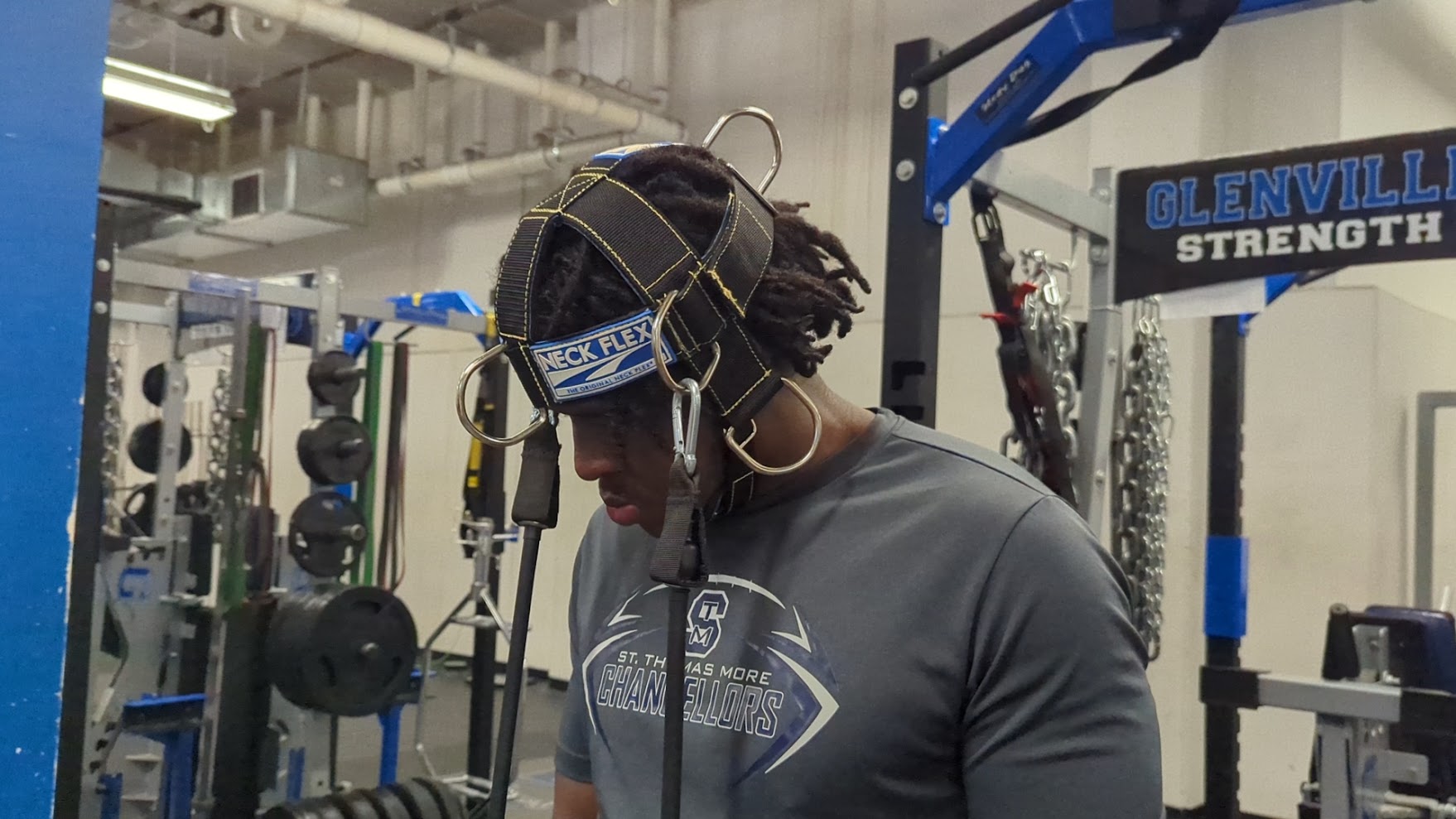 High school football player wearing a Neck Flex MMB Pro Series neck harness with band resistance during a team strength session in the Glenville Strength weight room
