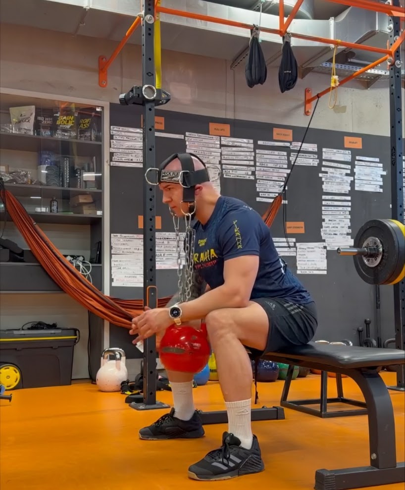 Lifter seated upright in a home gym wearing a Neck Flex harness loaded with a kettlebell on a chain while holding a resistance band anchored to the rack