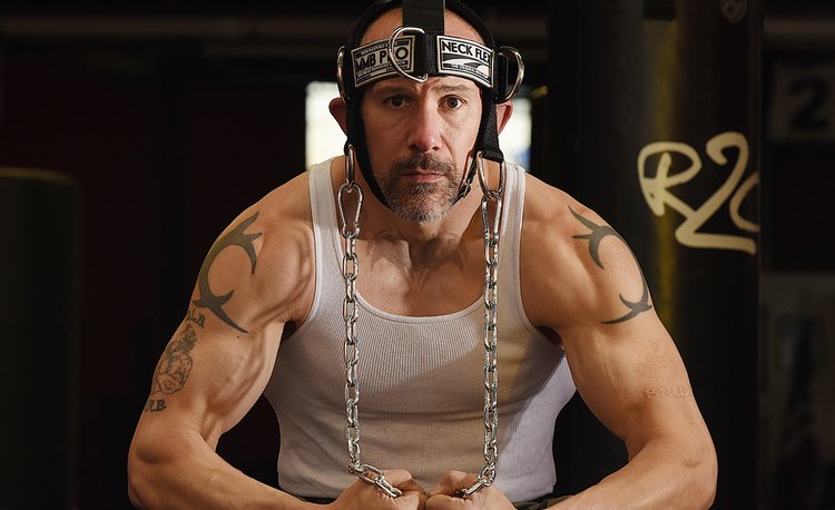 Man with tattoos wearing a white tank top and helmet with chains, in a dark setting.